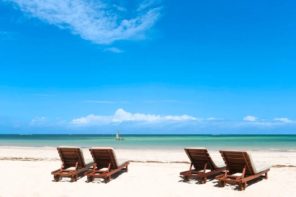 Quattro sedie a sdraio in legno costeggiano la spiaggia sabbiosa del Neptune Paradise Beach Resort, in Kenya, di fronte a un mare calmo e turchese con una barca a vela in lontananza, sotto un cielo blu brillante punteggiato di nuvole.