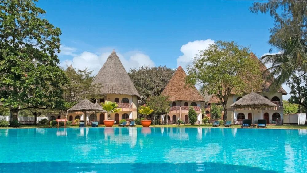 Una grande piscina trasparente circondata da palme e piante tropicali al Neptune Paradise Beach Resort Kenya, con edifici rotondi dai tetti di paglia sullo sfondo, sotto un cielo blu brillante.