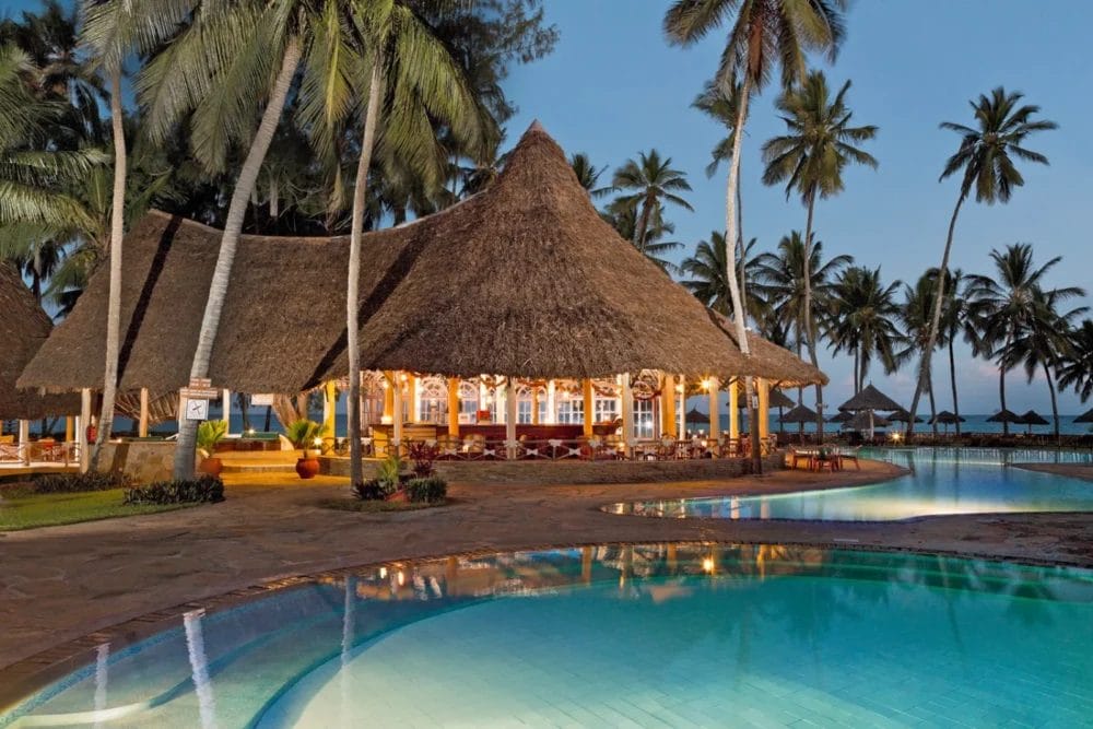 Un paradiso tropicale, il Neptune Paradise Beach Resort in Kenya è caratterizzato da un grande padiglione con tetto in paglia, circondato da palme e da una piscina trasparente al tramonto, con luci soffuse e un'invitante spa che crea un'atmosfera rilassante.