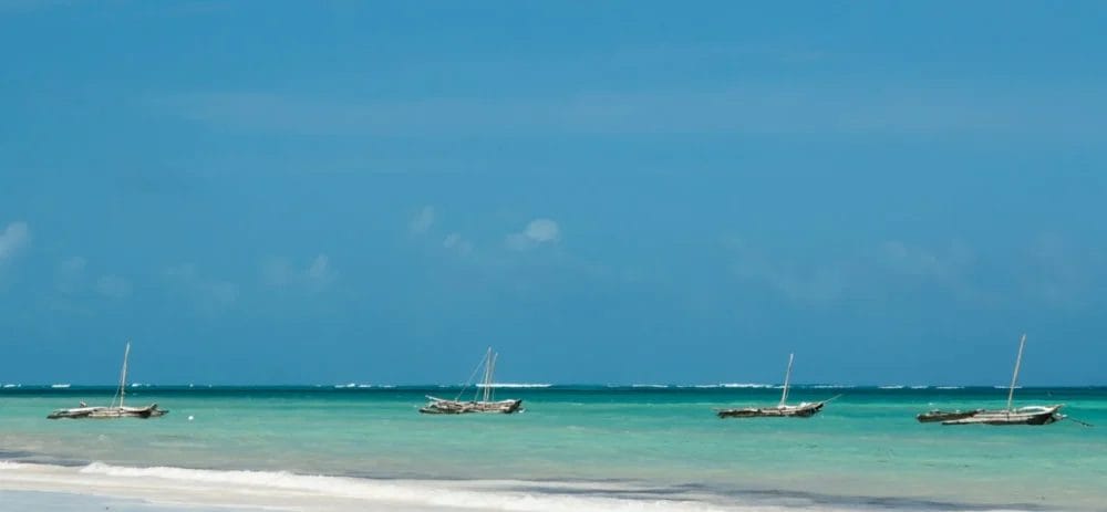 Quattro barche di legno galleggiano sull'acqua calma e turchese dell'oceano, vicino a una spiaggia bianca e sabbiosa del Neptune Paradise Beach Resort. La scena è pacifica e tropicale e cattura la serena bellezza di questo rinomato resort balneare in Kenya.