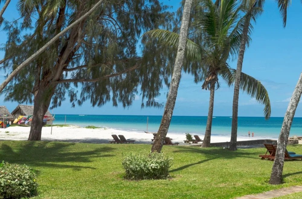 Palme e alberi frondosi incorniciano una spiaggia sabbiosa con acque turchesi dell'oceano al Neptune Paradise Beach Resort. Le sedie a sdraio sull'erba invitano al relax, mentre alcune persone passeggiano vicino alla riva sotto il cielo azzurro del Kenya.