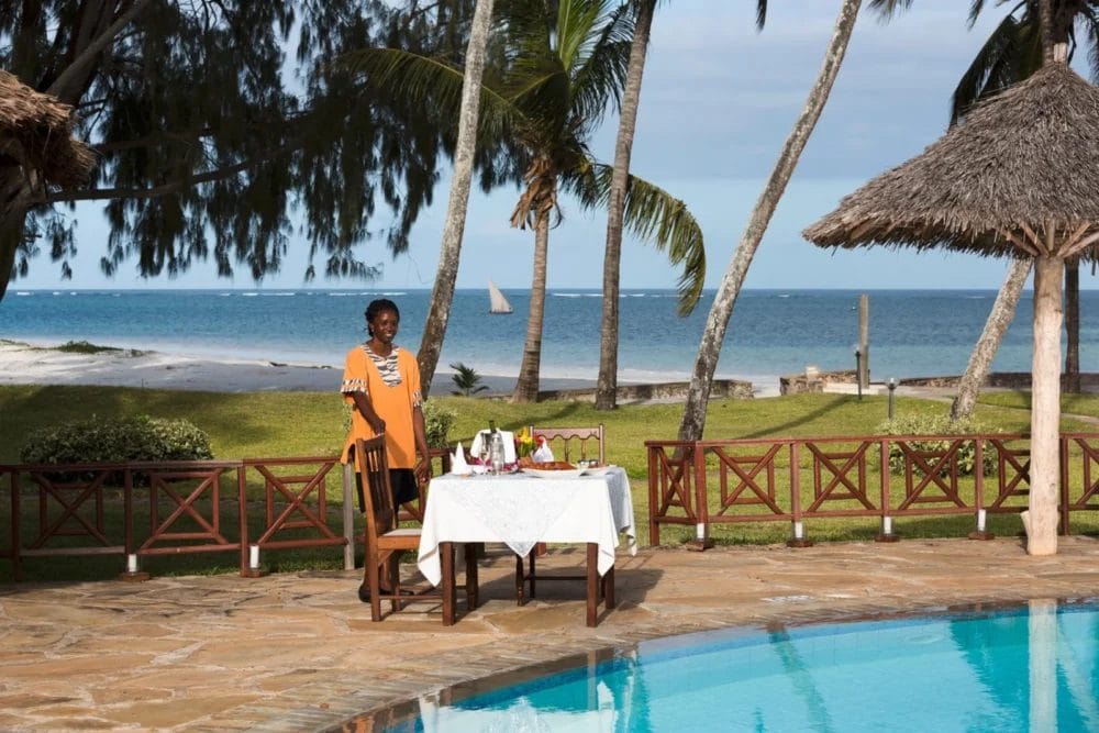Una persona in camicia arancione si trova accanto a un tavolo per la colazione apparecchiato per due persone, accanto alla piscina del Neptune Paradise Beach Resort, con palme, un'area erbosa e l'oceano sullo sfondo.
