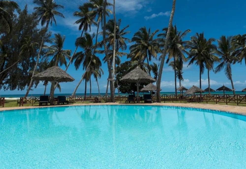 Una piscina azzurra con ombrelloni e sedie a sdraio di paglia, circondata da palme al Neptune Paradise Beach Resort, si affaccia su una spiaggia luminosa e soleggiata e sull'oceano sotto un cielo azzurro: la vostra fuga perfetta dal Kenya.