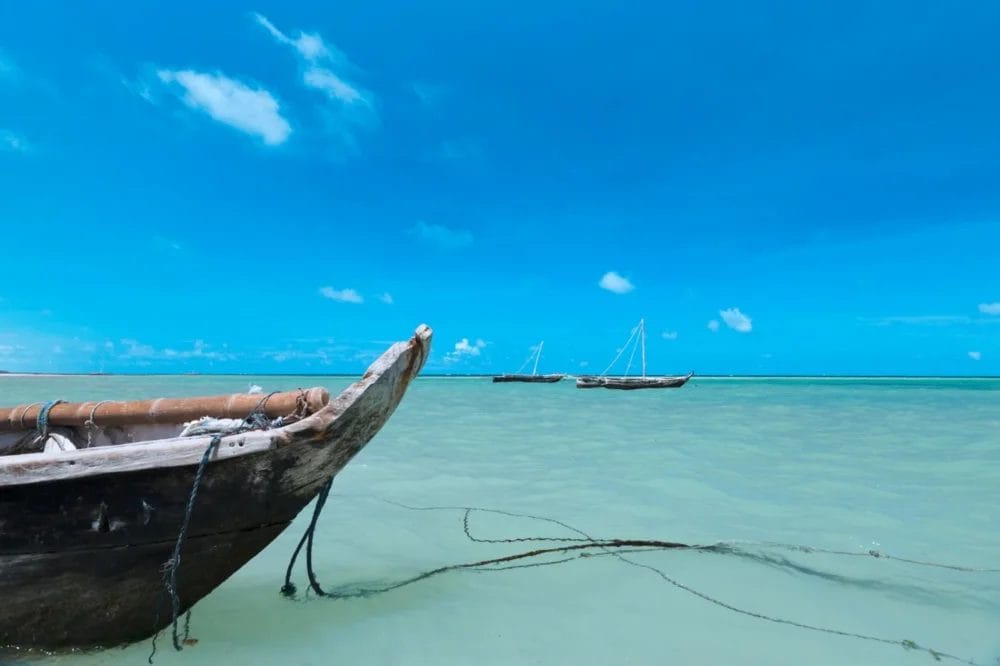 Una barca di legno è ancorata in acque turchesi poco profonde vicino al Neptune Paradise Beach Resort, con altre due barche che galleggiano in lontananza sotto un cielo blu brillante disseminato di piccole nuvole.