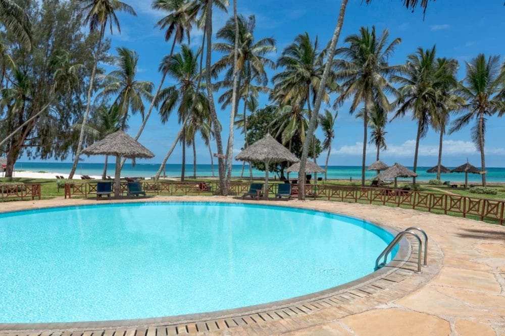 Una piscina rotonda con acqua cristallina è circondata da palme, recinzioni in legno e ombrelloni di paglia al Neptune Paradise Beach Resort. Oltre la piscina si trova una spiaggia sabbiosa e l'oceano sotto il cielo luminoso e soleggiato del Kenya.