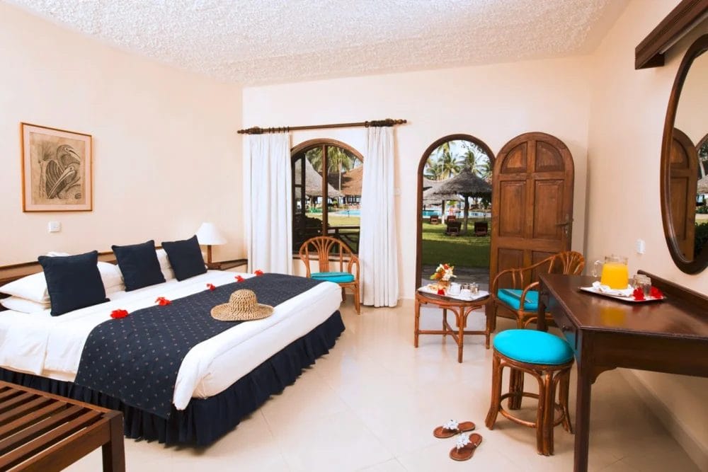 Una luminosa camera d'albergo con letto matrimoniale, biancheria da letto blu e cuscini decorativi al Neptune Paradise Beach Resort. Due sedie, un tavolino con bevande e frutta e grandi finestre ad arco si affacciano su giardini lussureggianti in questo rilassante resort sulla spiaggia in Kenya.