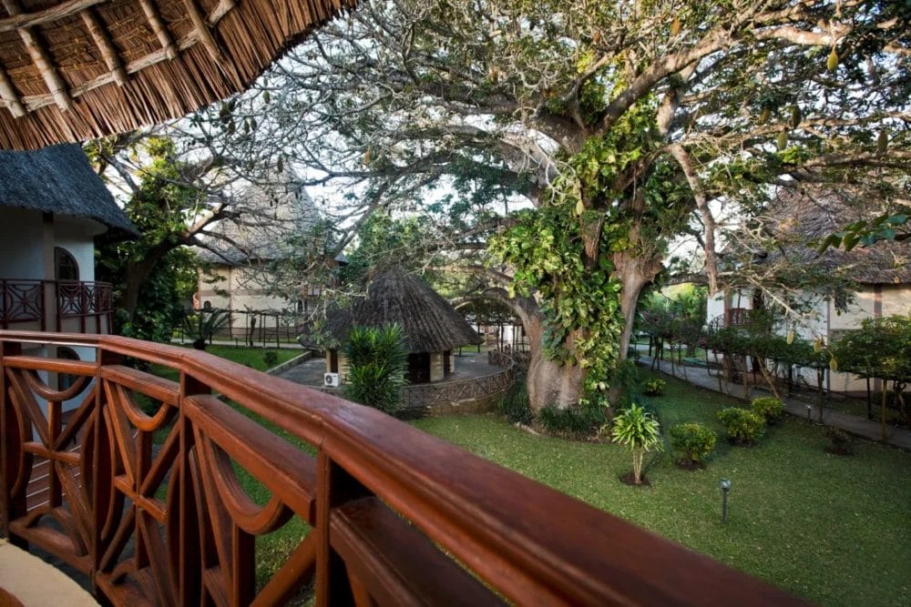 Una vista da un balcone con ringhiere in legno che si affaccia su un giardino verdeggiante, grandi alberi e strutture con tetto in paglia nella tranquilla cornice esterna del Neptune Paradise Beach Resort, un rilassante resort sulla spiaggia in Kenya.