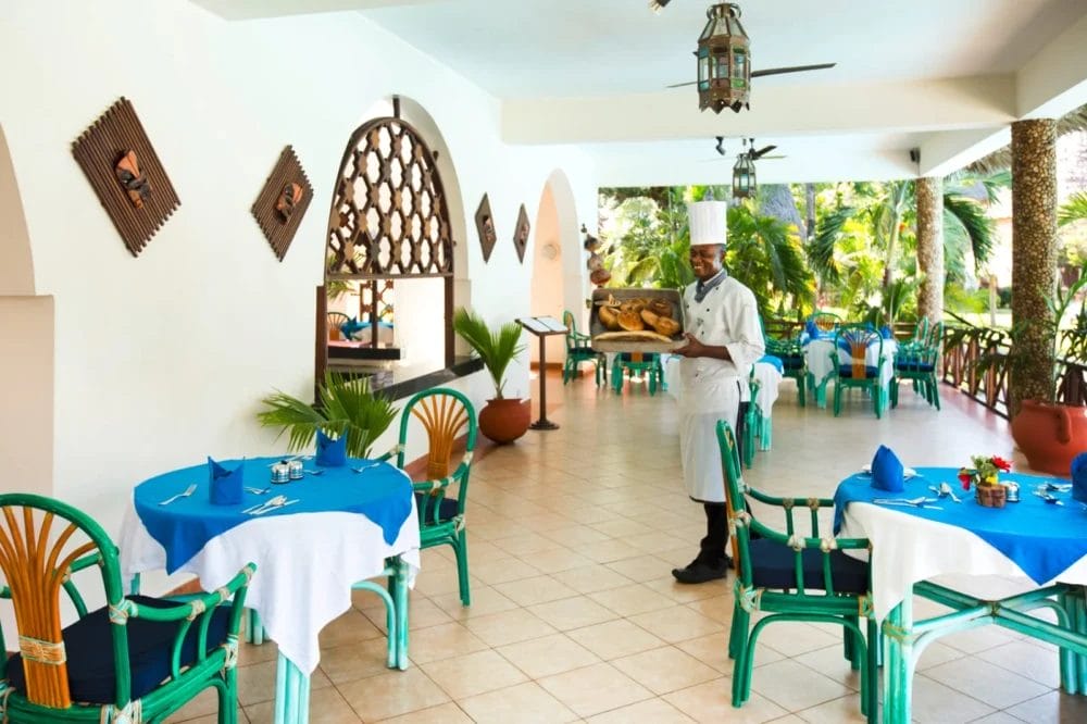Uno chef in uniforme e cappello bianco si trova nel ristorante all'aperto del Neptune Paradise Beach Resort, con in mano un vassoio di pane. Lo spazio luminoso è caratterizzato da sedie verdi, tovaglie blu, piante tropicali e archi che si aprono su un giardino lussureggiante.