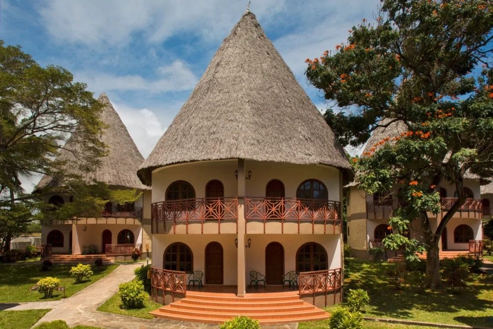 Edificio rotondo con un alto tetto conico di paglia, due livelli di balconi con ringhiere in legno, immerso tra alberi verdi e giardini paesaggistici del Neptune Paradise Beach Resort sotto un cielo blu brillante.