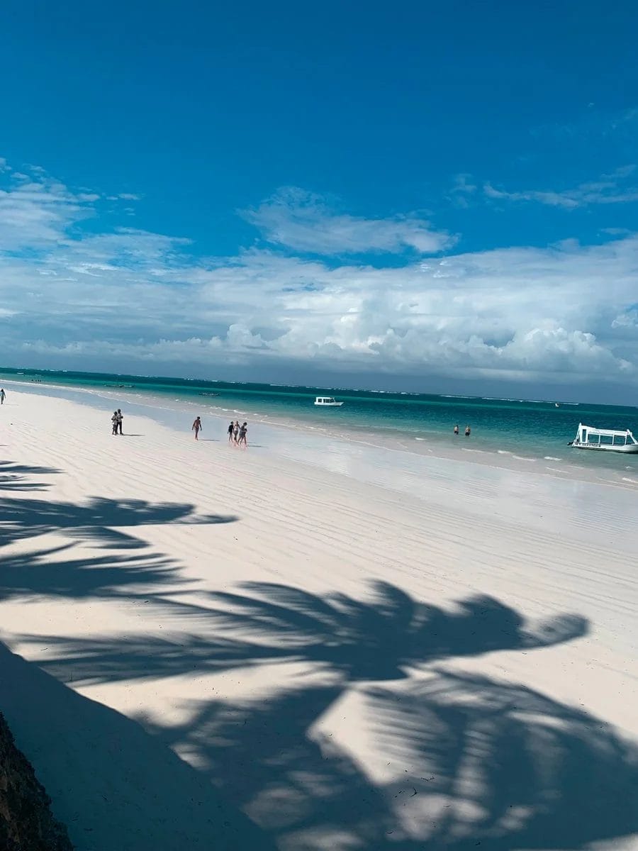 Una scena di sole sulla spiaggia con sabbia bianca, acqua turchese e cielo azzurro al Neptune Paradise Beach Resort & Spa Kenya. Alcune persone camminano lungo la riva, due barche galleggiano sull'acqua e le ombre delle palme si allungano sulla sabbia.