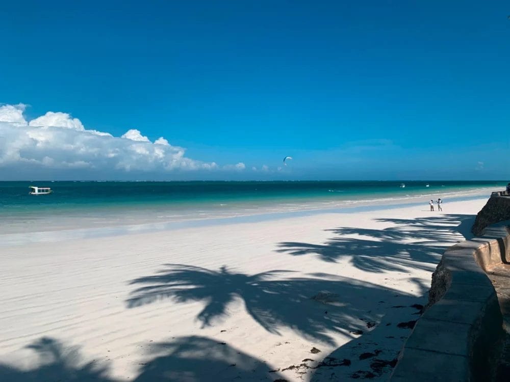 Una spiaggia soleggiata con sabbia bianca, acqua turchese e cielo azzurro presso il Neptune Paradise Beach Resort in Kenya. Le ombre delle palme si allungano sulla sabbia, mentre alcune persone e una barca sono visibili in lontananza.