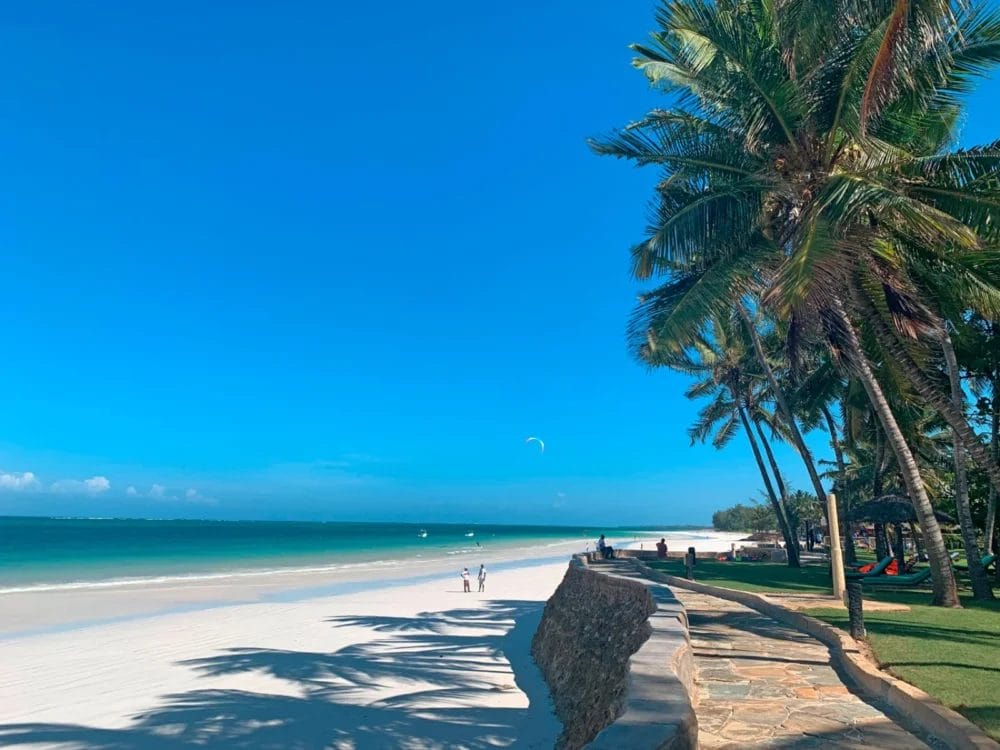 Una spiaggia soleggiata con sabbia bianca, acqua turchese e cielo azzurro. Le palme si appoggiano a un muro di pietra sulla destra, mentre le persone si rilassano lungo la riva vicino al Neptune Paradise Beach Resort, uno dei migliori resort balneari del Kenya.