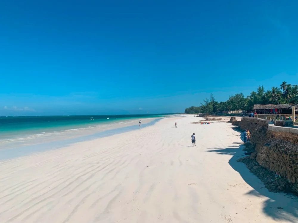 Un'ampia spiaggia sabbiosa con acqua turchese sotto un cielo azzurro e limpido. Alcune persone passeggiano lungo la riva vicino al Neptune Paradise Beach Resort, in Kenya, con alberi e strutture termali visibili sul lato destro dell'immagine.