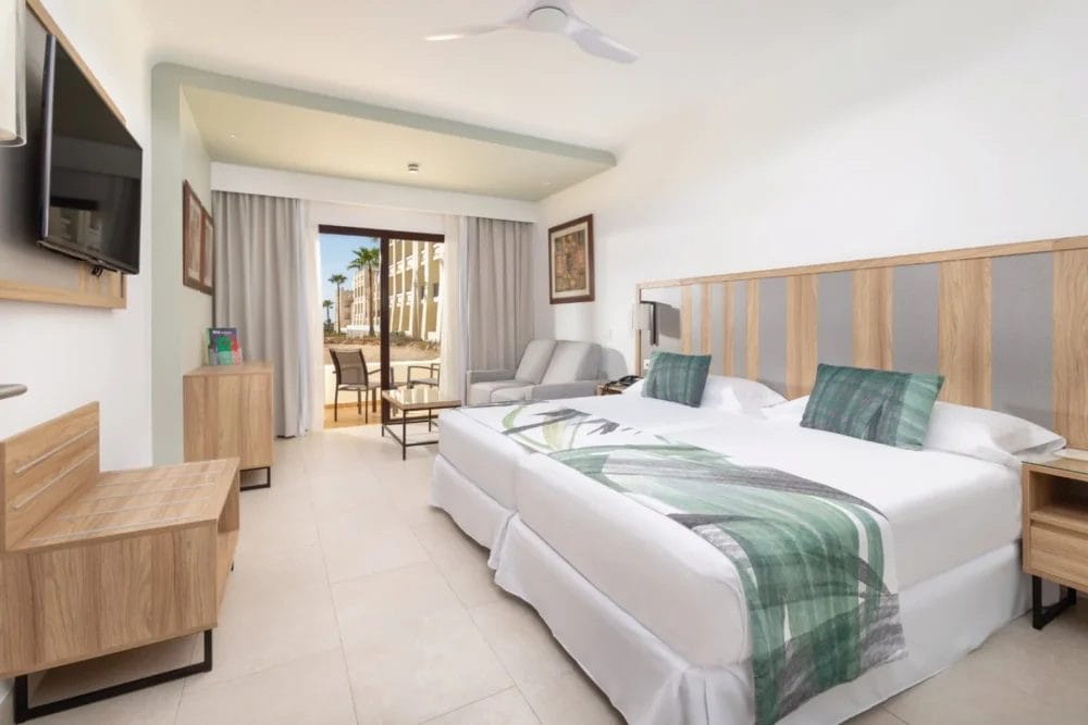Moderna camera d'albergo SEARESORT con due letti singoli accostati, cuscini verdi, TV a parete, mobili in legno chiaro, piccola area salotto e porta a vetri che conduce a un patio esterno con palme al RIU KARAMBOA BOAVISTA.
