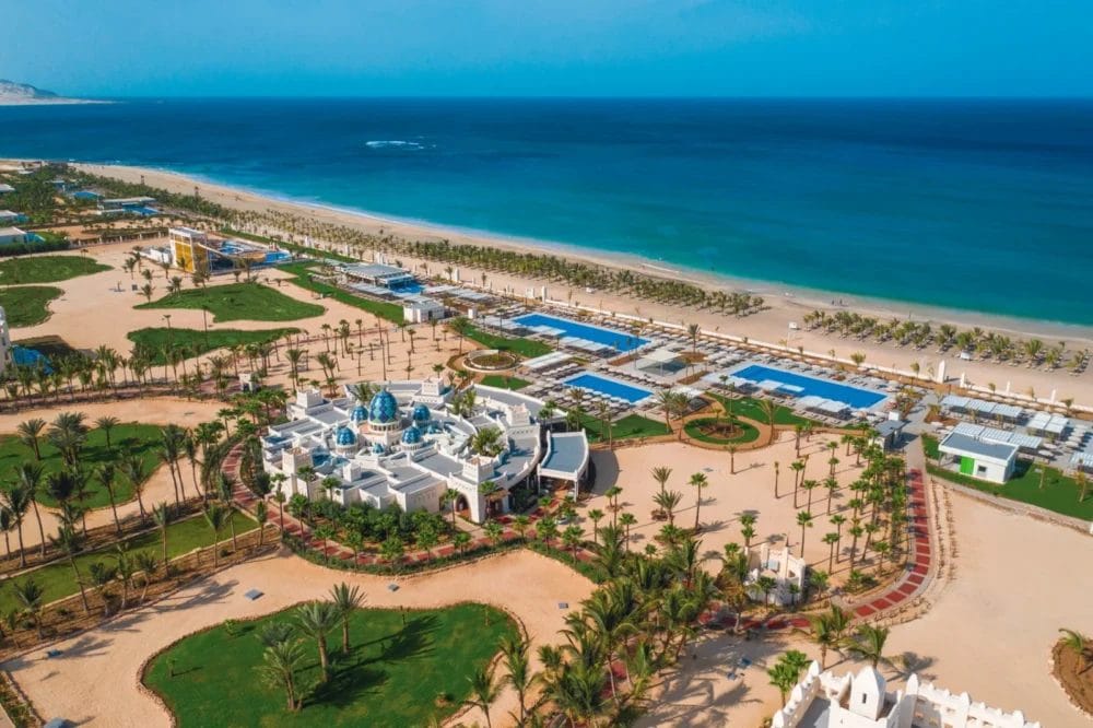 Vista aerea del SEARESORT RIU KARAMBOA, un resort sulla spiaggia di BOAVISTA con edifici bianchi, cupole blu, palme e prati verdi lungo una spiaggia sabbiosa e l'oceano blu che brilla sotto un cielo limpido.