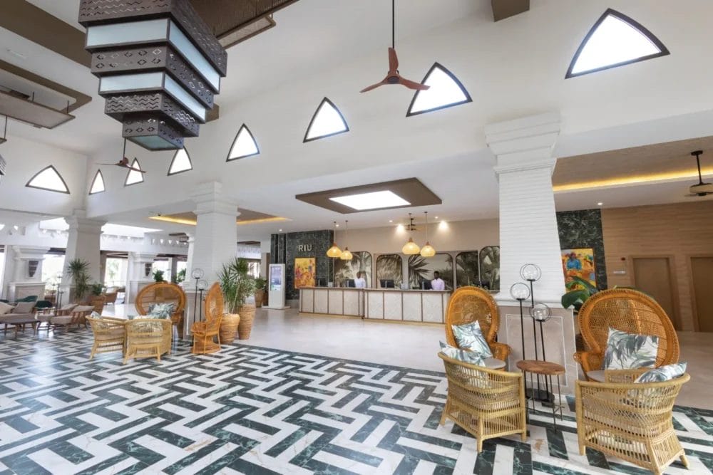 La spaziosa hall del RIU KARAMBOA on BOAVISTA è caratterizzata da un pavimento in piastrelle con motivi a zig zag, sedie in vimini con cuscini, colonne bianche e un soffitto alto. Le grandi finestre e l'arredamento moderno creano un'atmosfera luminosa e ariosa da SEARESORT.