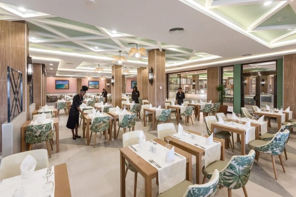 Un ristorante moderno al RIU KARAMBOA con tavoli in legno apparecchiati per i pasti, sedie verdi e bianche imbottite, luci decorative sul soffitto e personale che prepara lo spazio. L'ambiente è luminoso e spazioso con arredamento contemporaneo SEARESORT in BOAVISTA.