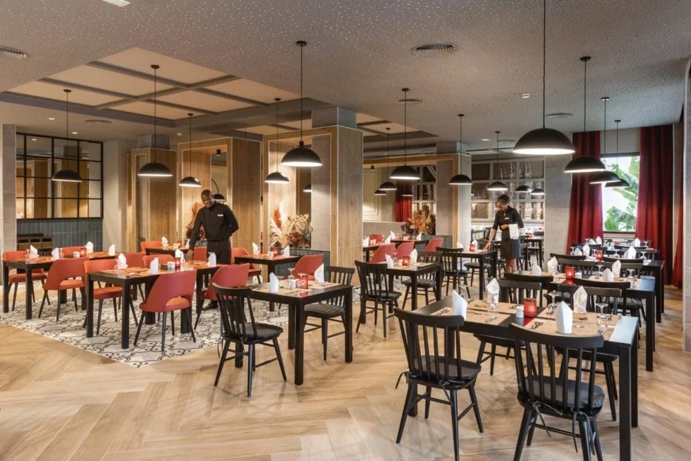 Ristorante moderno del SEARESORT BOAVISTA con pavimenti in legno, sedie rosse e nere, tavoli ordinati e luci a sospensione. Due membri del personale che indossano maschere si preparano per il servizio nell'atmosfera calda e invitante del RIU KARAMBOA.