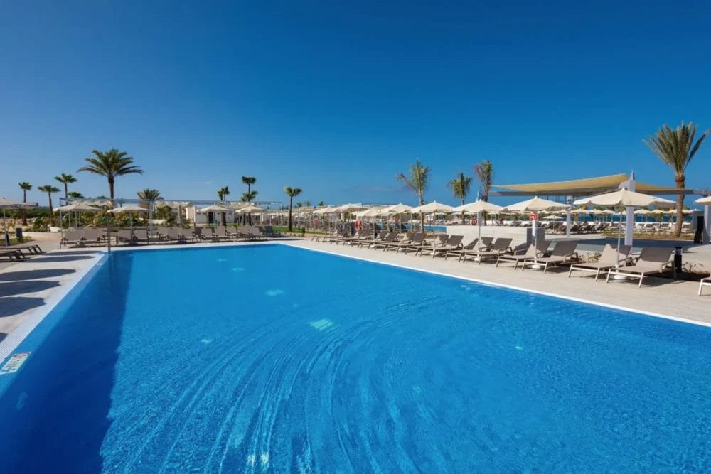 La grande piscina blu chiaro di SEARESORT è circondata da sedie a sdraio e ombrelloni, con palme e un cielo azzurro brillante sullo sfondo. Lo scenario soleggiato di BOAVISTA è davvero invitante.