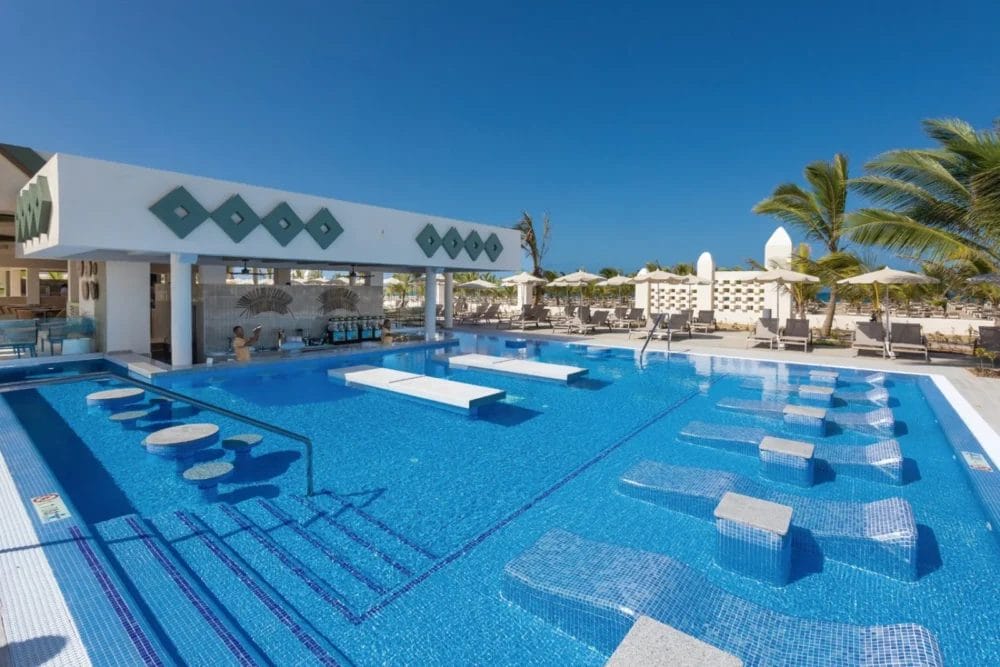 La moderna piscina del SEARESORT RIU KARAMBOA a Boavista è dotata di sedie a sdraio sommerse, bar in acqua e bar a bordo piscina. Palme, ombrelloni e sedie a sdraio circondano la piscina sotto un cielo azzurro e limpido.