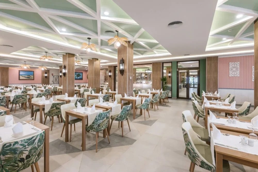Gli interni del ristorante del RIU KARAMBOA, spaziosi e moderni, con tavoli di legno ordinati, sedie verdi a fantasia, coperti bianchi, luci a sospensione e pannelli decorativi sul soffitto, creano un'atmosfera luminosa e invitante.