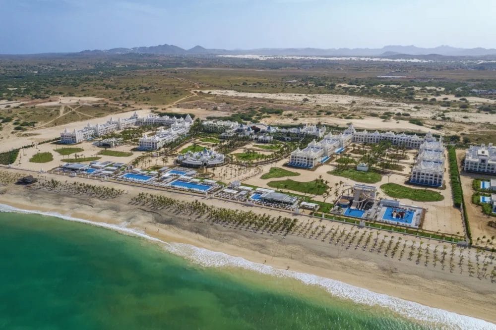 Vista aerea del RIU Karamboa, uno splendido resort sul mare a Boavista, con edifici bianchi, piscine blu, palme e spiaggia sabbiosa sullo sfondo di un paesaggio arido e di montagne lontane.