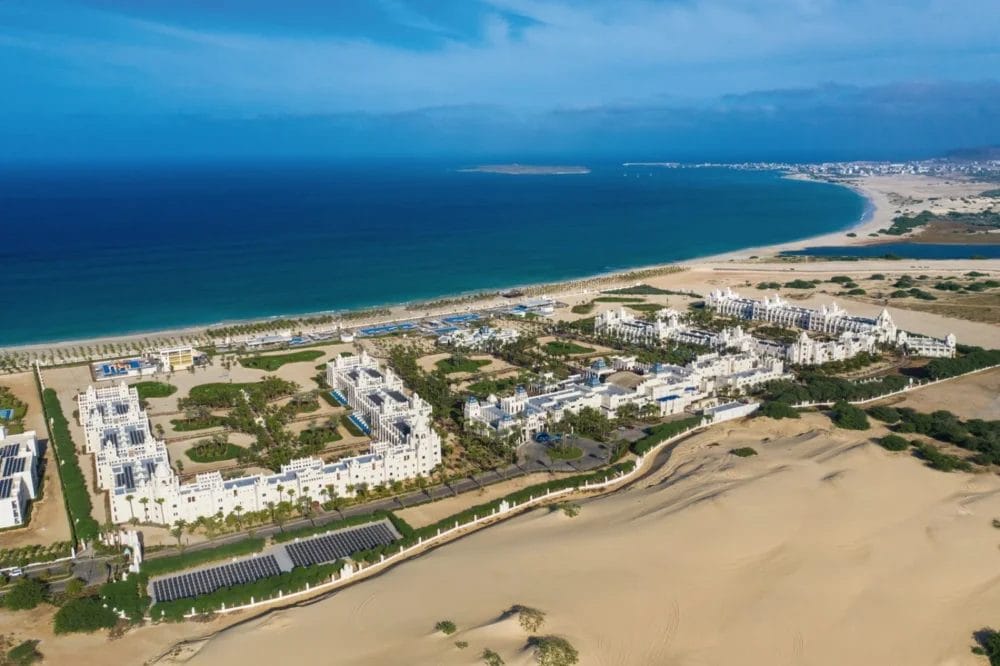 Vista aerea del SEARESORT RIU KARAMBOA, un grande complesso di resort bianchi a Boavista, circondato da verde e dune di sabbia accanto a una lunga spiaggia sinuosa con oceano blu, sotto un cielo parzialmente nuvoloso.