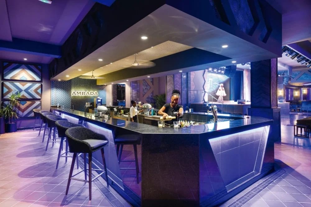 Un moderno bar del RIU Karamboa sea resort con un design geometrico, piani d'appoggio scuri, sgabelli da bar e un'illuminazione ambientale viola. Un barista prepara i drink mentre una cantante si esibisce sul palco di Boavista. Sul muro compare l'insegna "Ambada".
