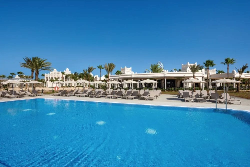Una grande piscina all'aperto con acqua cristallina al SEARESORT RIU KARAMBOA, circondata da sedie a sdraio e ombrelloni. Le palme e l'edificio bianco del resort Boavista si stagliano sotto un cielo azzurro e limpido.