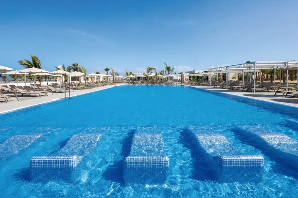 Una grande piscina all'aperto del RIU KARAMBOA con sedie a sdraio sommerse, circondata da lettini e ombrelloni, palme e un cielo azzurro e limpido di BOAVISTA.
