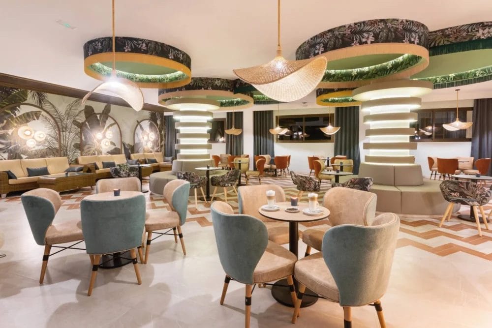 Moderna lounge del SEARESORT Boavista con morbide sedie beige e verde acqua attorno a piccoli tavoli, singolari luci circolari sul soffitto e decorazioni contemporanee, tra cui grandi colonne e opere d'arte a parete a forma di foglie. Lo spazio è ben illuminato e arredato con stile.