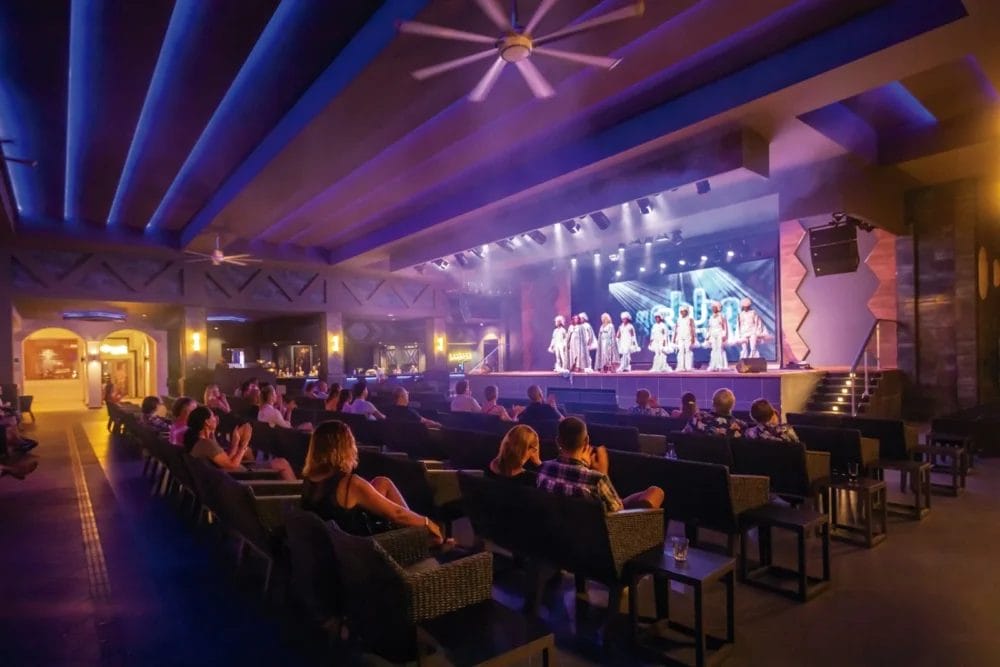 Un gruppo di artisti si trova su un palcoscenico illuminato di fronte a un pubblico nel moderno teatro coperto RIU KARAMBOA, le cui luci blu e i ventilatori a soffitto evocano l'atmosfera vibrante del SEARESORT di BOAVISTA.