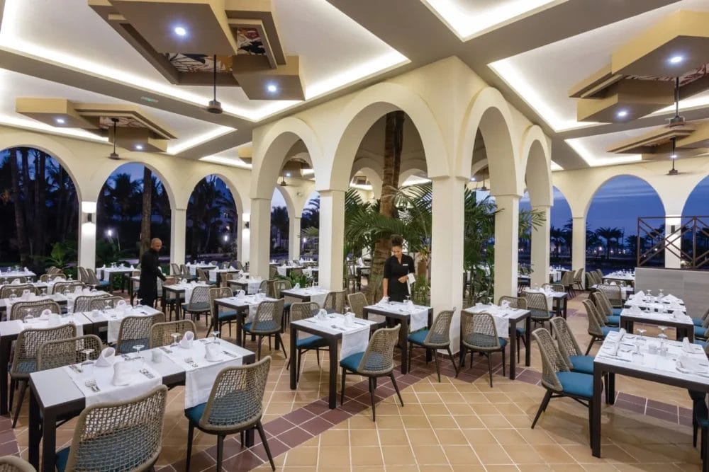 Elegante ristorante del RIU KARAMBOA con colonne bianche ad arco, moderne luci a soffitto e tavoli ordinati. Due addetti preparano lo spazio mentre le grandi finestre rivelano palme e un cielo serale sul SEARESORT di BOAVISTA.
