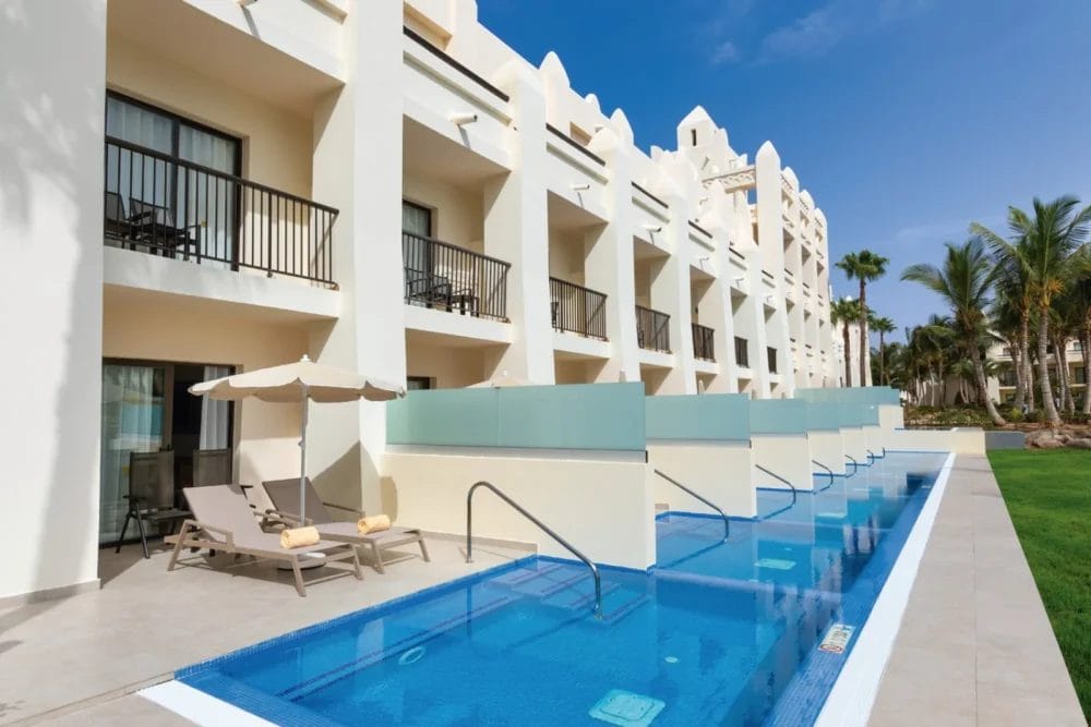 Una fila di moderne camere dell'hotel SEARESORT con balcone privato si affaccia sulla lunga e stretta piscina di Boavista. Ogni camera ha accesso diretto alla piscina, sedie a sdraio e ombrelloni. Palme ed erba verde sono incastonati sotto il cielo blu.