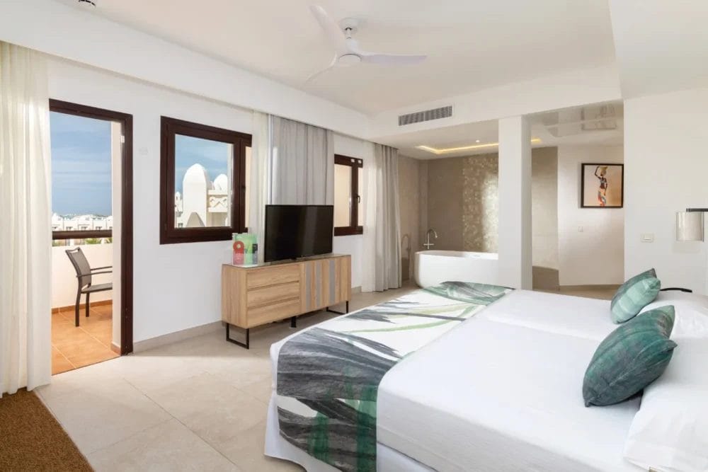 Moderna camera d'albergo del SEARESORT RIU KARAMBOA con letto matrimoniale con lenzuola verdi e bianche, TV a schermo piatto su un supporto in legno, ampie finestre con tende, balcone e area bagno aperta con vasca da bagno sullo sfondo, perfetta per la vostra fuga a BOAVISTA.