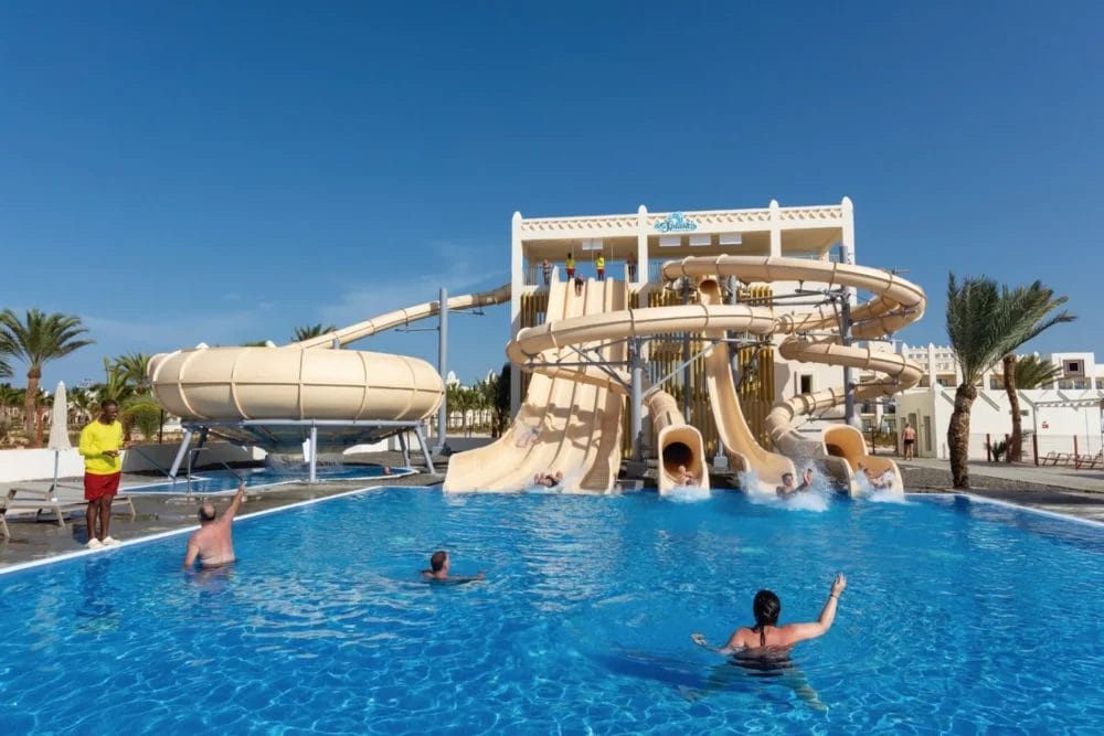 Le persone si godono una giornata di sole al parco acquatico RIU Karamboa Sea Resort di Boavista, con una grande piscina blu e scivoli abbronzanti. Alcuni scivolano in piscina mentre altri nuotano o si rilassano, circondati da palme e da un cielo azzurro e limpido.