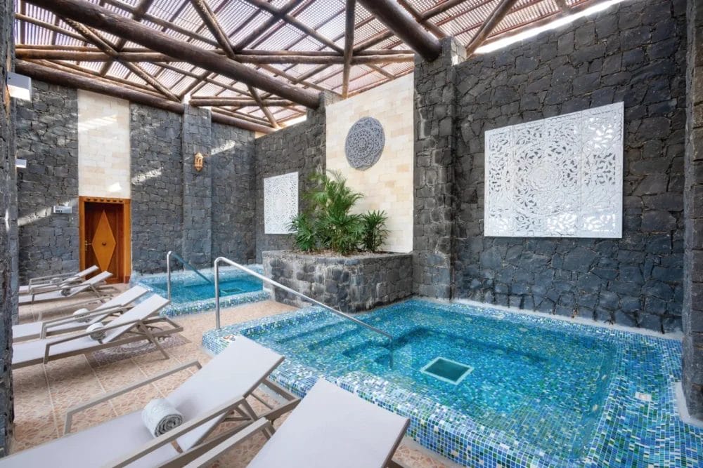 Area spa interna del RIU Karamboa, con piccole piscine in piastrelle blu, pareti in pietra, pannelli decorativi bianchi, sedie a sdraio con asciugamani, piante tropicali e una struttura in legno sul tetto che lascia entrare la luce naturale: un rifugio sereno in questo resort sul mare di Boavista.