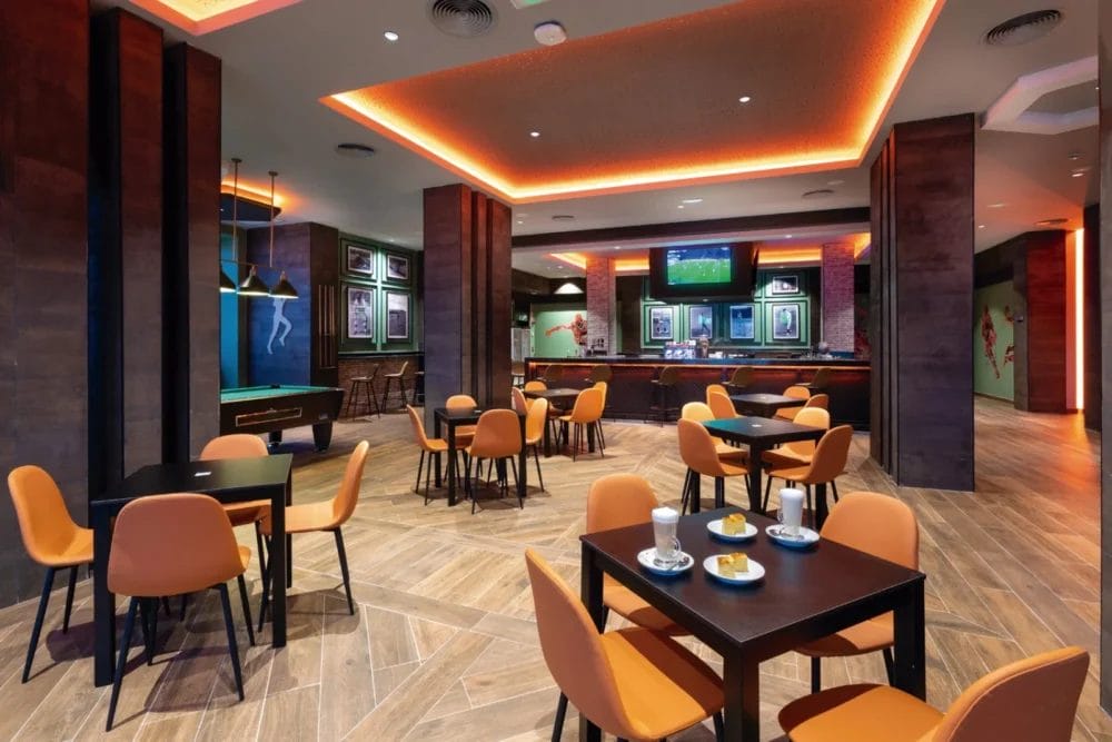 Un moderno salone del SEARESORT Boavista con sedie arancioni e tavoli scuri, apparecchiati con bevande e snack. Sono visibili un tavolo da biliardo, un'area bar e diversi televisori, mentre le calde luci del soffitto creano un'accogliente atmosfera da RIU KARAMBOA.