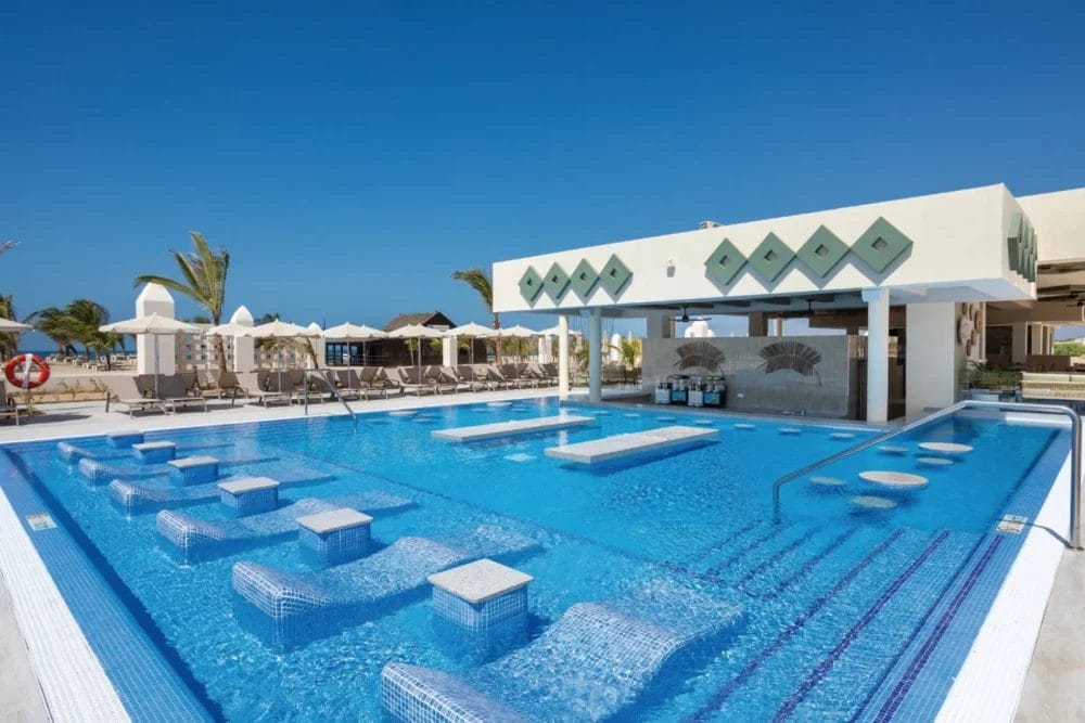 Un moderno bar a bordo piscina con sgabelli sommersi in una piscina blu chiaro al SEARESORT RIU KARAMBOA, Boavista. Sedie a sdraio e ombrelloni costeggiano il bordo della piscina, mentre le palme e la spiaggia sono visibili sullo sfondo.
