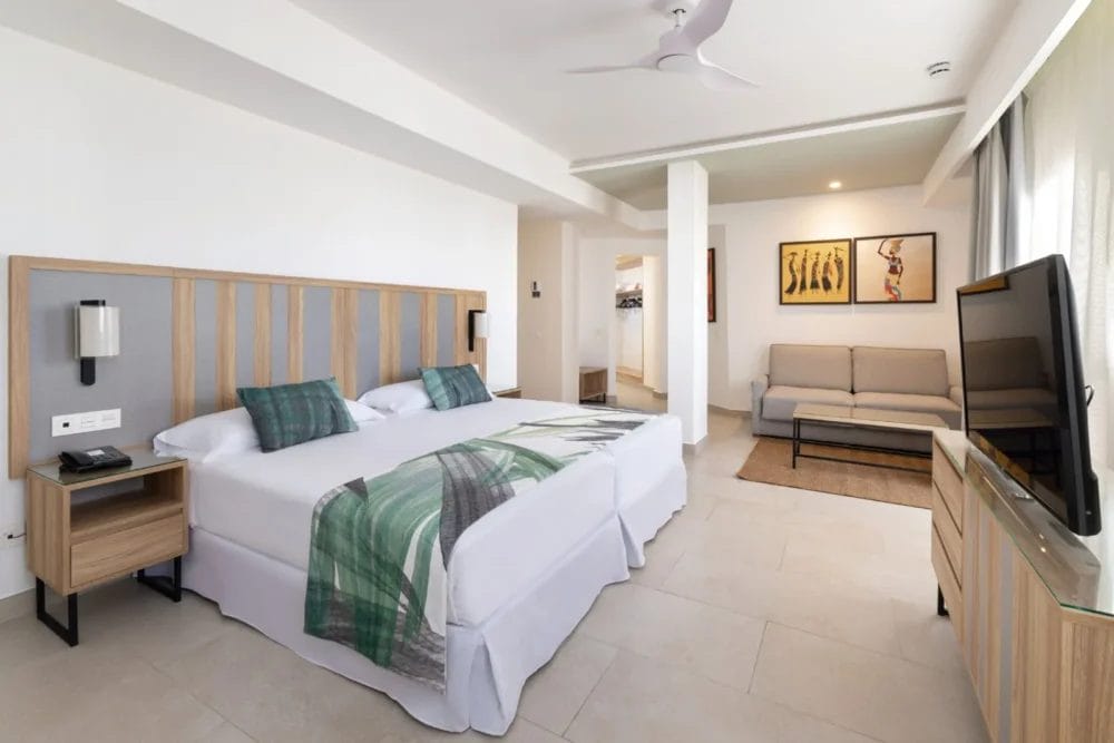 Moderna camera d'albergo al RIU KARAMBOA con due letti singoli accostati, coperte verdi a fantasia, comodini, divano, opere d'arte, TV su un mobile, ventilatore a soffitto e ampia finestra con tende trasparenti che lasciano entrare la luce naturale.