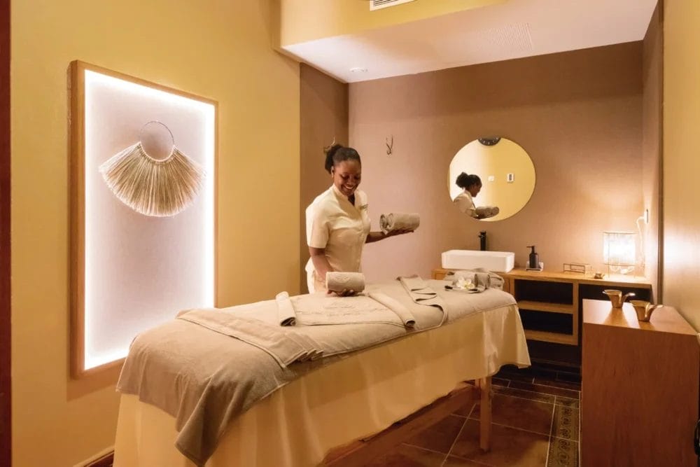 Un massaggiatore sorridente si trova accanto a un lettino per massaggi preparato con cura in una sala spa del SEARESORT RIU KARAMBOA, illuminata da luci soffuse e decorata con colori caldi e opere d'arte minimaliste.