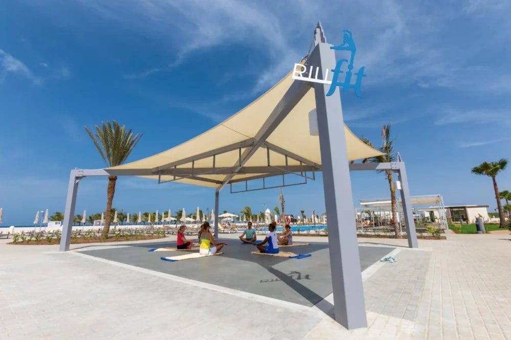 Un gruppo pratica yoga su tappetini sotto un grande e moderno ombrellone con la scritta "Blu Fit" al RIU Karamboa SEARESORT, con le palme e la spiaggia di Boavista visibili sullo sfondo soleggiato.
