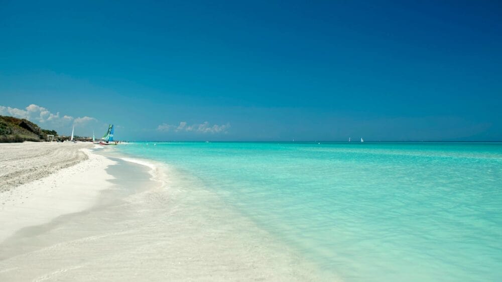 Una spiaggia incontaminata con sabbia bianca e acqua limpida e turchese, delimitata da un mare calmo sotto un cielo azzurro brillante. Alcune barche e velieri alla deriva vicino alla riva, evocano ricordi di Jibacoa, tra L'Avana e Varadero, vicino a incantevoli case particulars.