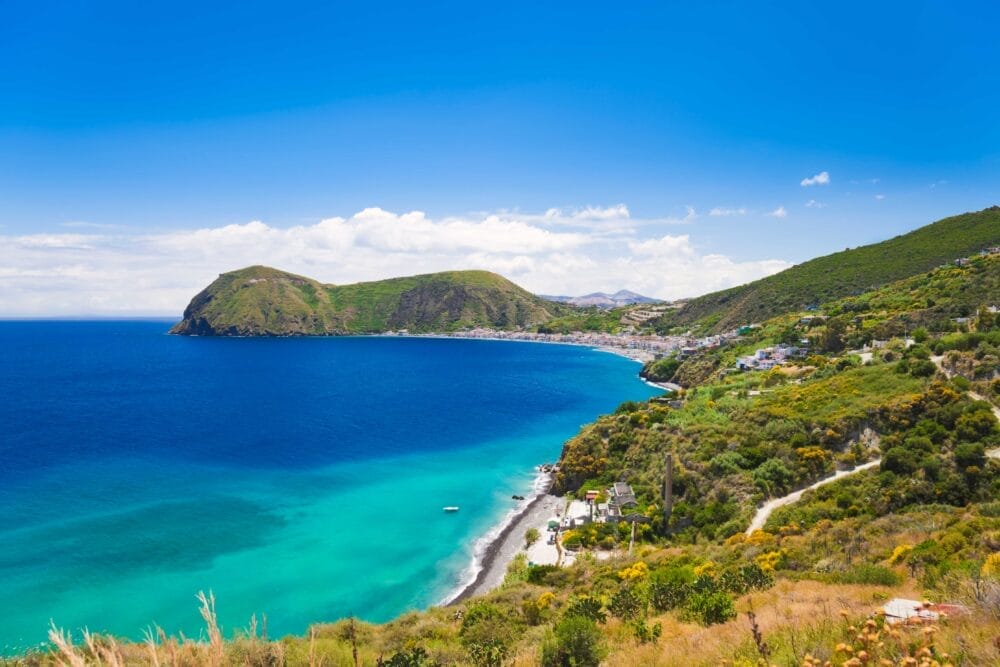 Una vista panoramica della costa sinuosa delle ISOLE EOLIE, con il mare blu turchese, le colline verdeggianti e un piccolo villaggio lungo la riva, sotto un cielo blu brillante con nuvole sparse: l'ideale per il vostro prossimo tour Eolie.