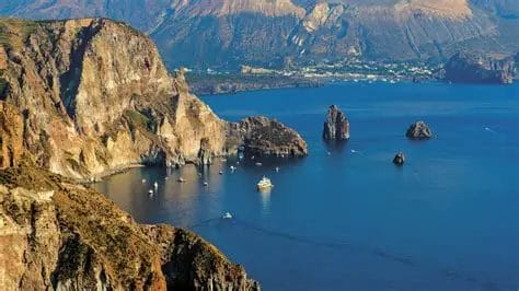 Una vista di scogliere rocciose e di faraglioni che emergono dall'acqua blu delle Isole Eolie, con diverse barche e una piccola città visibili lungo la costa: uno scenario perfetto per il vostro prossimo viaggio o tour panoramico. Le aspre montagne completano la scena.