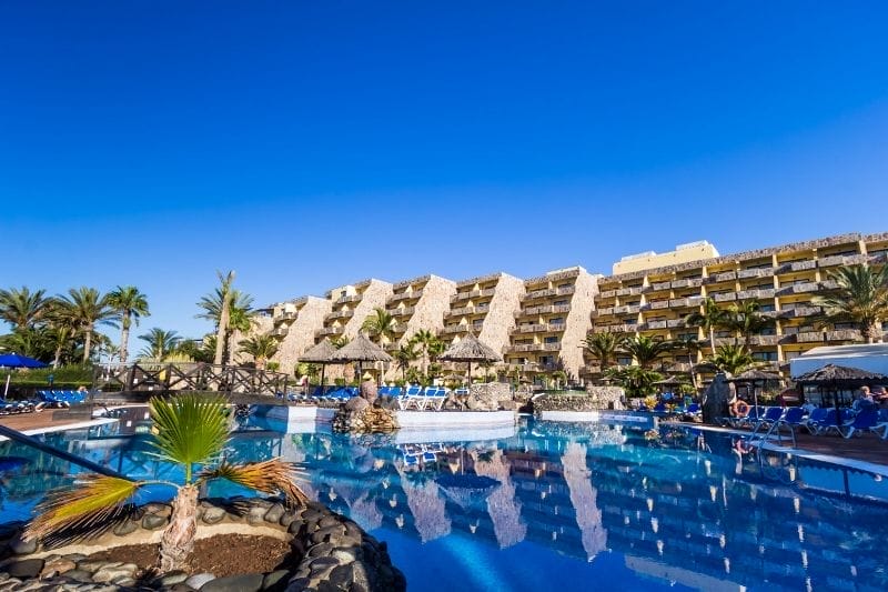 Un resort con edifici a terrazze e palme si riflette in una grande piscina all'aperto sotto un cielo azzurro e limpido. Sedie a sdraio e ombrelloni costeggiano il bordo della piscina dell'HOTEL BLUEBAY, un hotel 4 stelle caratterizzato da un'esclusiva atmosfera da BEACH CLUB.