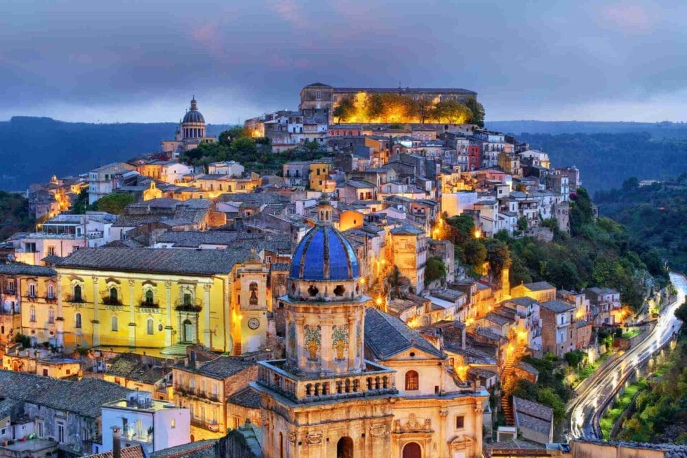 Una pittoresca città collinare al tramonto con edifici colorati, strade strette e luci scintillanti. Perfetta per un tour della Sicilia orientale, la scena presenta una chiesa dalle cupole blu in primo piano, tra il verde lussureggiante e le dolci colline.