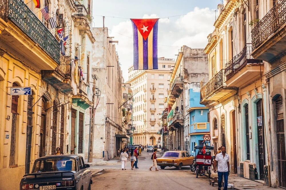 Una vivace scena di strada a L'Avana, Cuba, con auto d'epoca, vecchi edifici colorati, persone che camminano, una grande bandiera cubana appesa sopra la strada e una vicina Casa Particular che offre un autentico soggiorno locale.