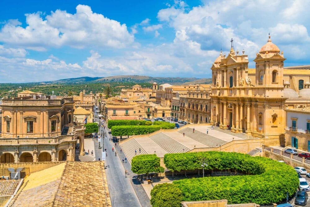 Una vista panoramica di Noto, in Sicilia, invita a visitare la Sicilia orientale, con edifici storici barocchi, una grande cattedrale con due campanili gemelli, scalinate in pietra, alberi frondosi e un cielo azzurro punteggiato di nuvole.