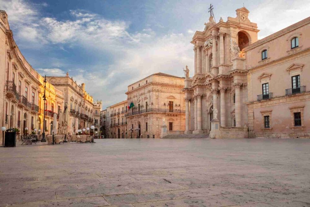 Un'ampia piazza in pietra delimitata da edifici storici barocchi, tra cui una grande cattedrale con colonne e statue, vi invita a esplorare la Sicilia orientale sotto un cielo azzurro parzialmente nuvoloso.